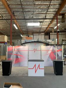 RENTAL: RE-2009 Inline Design with Arch Canopy with Black Pillowcase Cover, (2) RE-1201 Black Tapered Workstation Counters, (1) RE-1202 Rectangular Reception Counter, (8) Small Clear Acrylic Shelves, (8) LED Arm Lights, Tension Fabric Graphics, Direct Pri